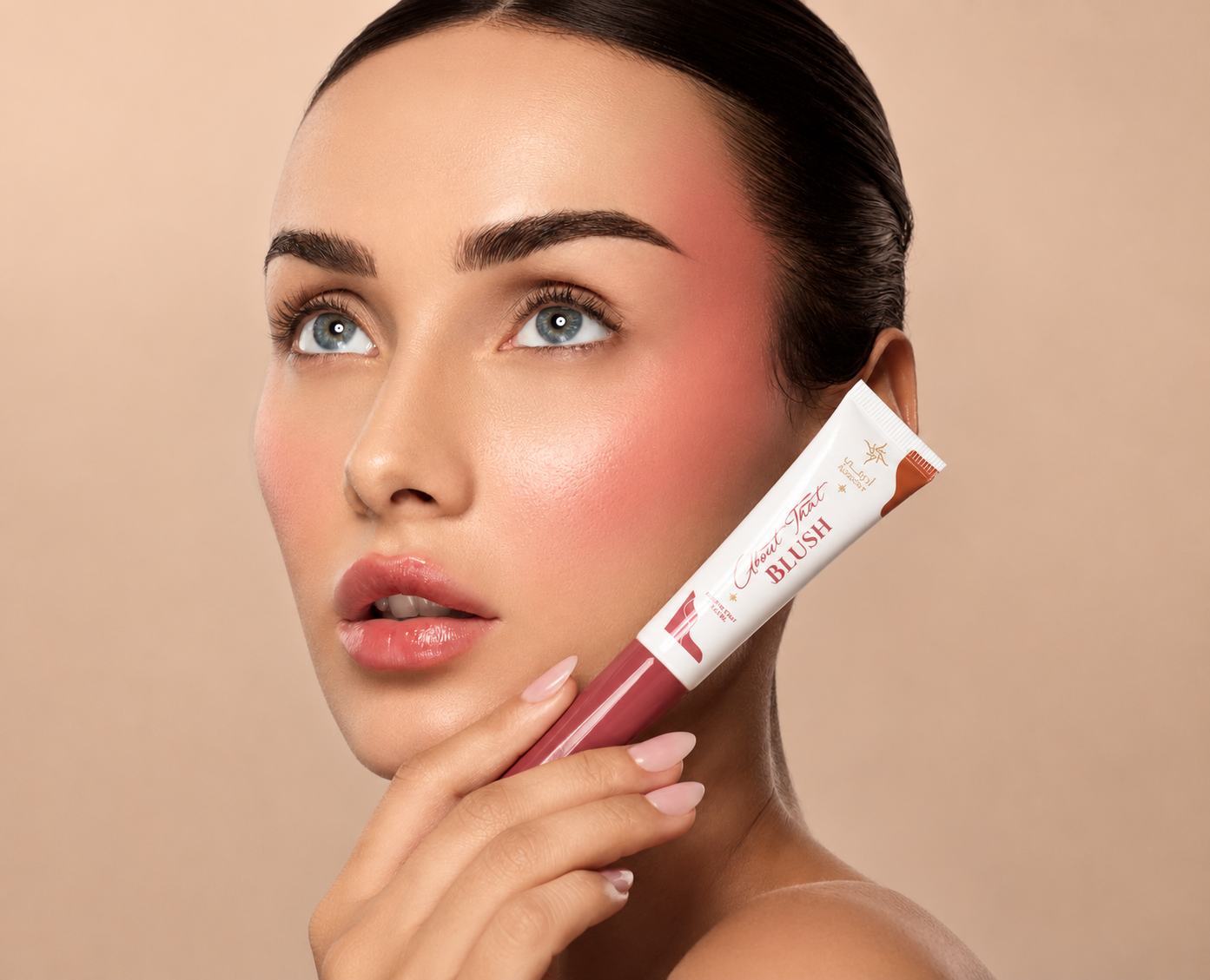 A close-up beauty shot of a woman holding a tube of blush against her cheek, with her face and hand visible against a soft beige background.