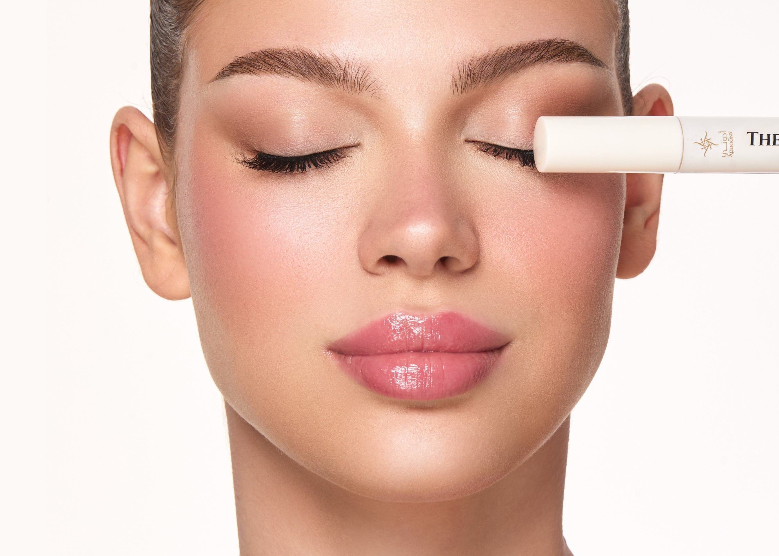 Close-up of a woman with closed eyes applying or showing a white cosmetic tube near her eye, on a clean white background.