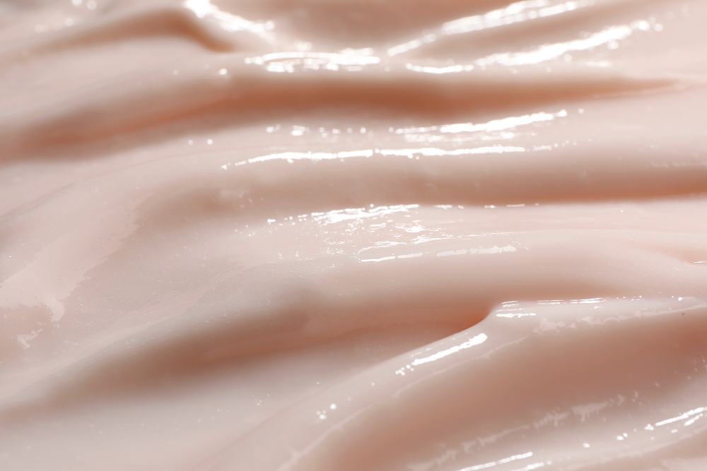 A close-up texture shot of a smooth, glossy, pale pink cream or lotion with soft ridges and highlights.