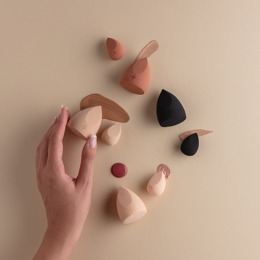 Hand holding a makeup sponge with various other sponges on a beige background