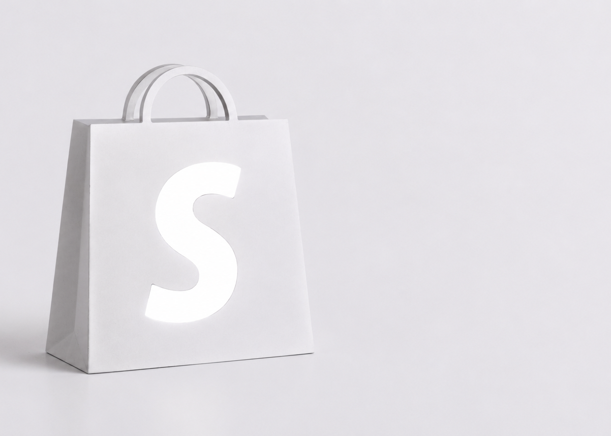 A minimalist white paper shopping bag with a large white letter 'S' on the front, set against a seamless light gray studio background.