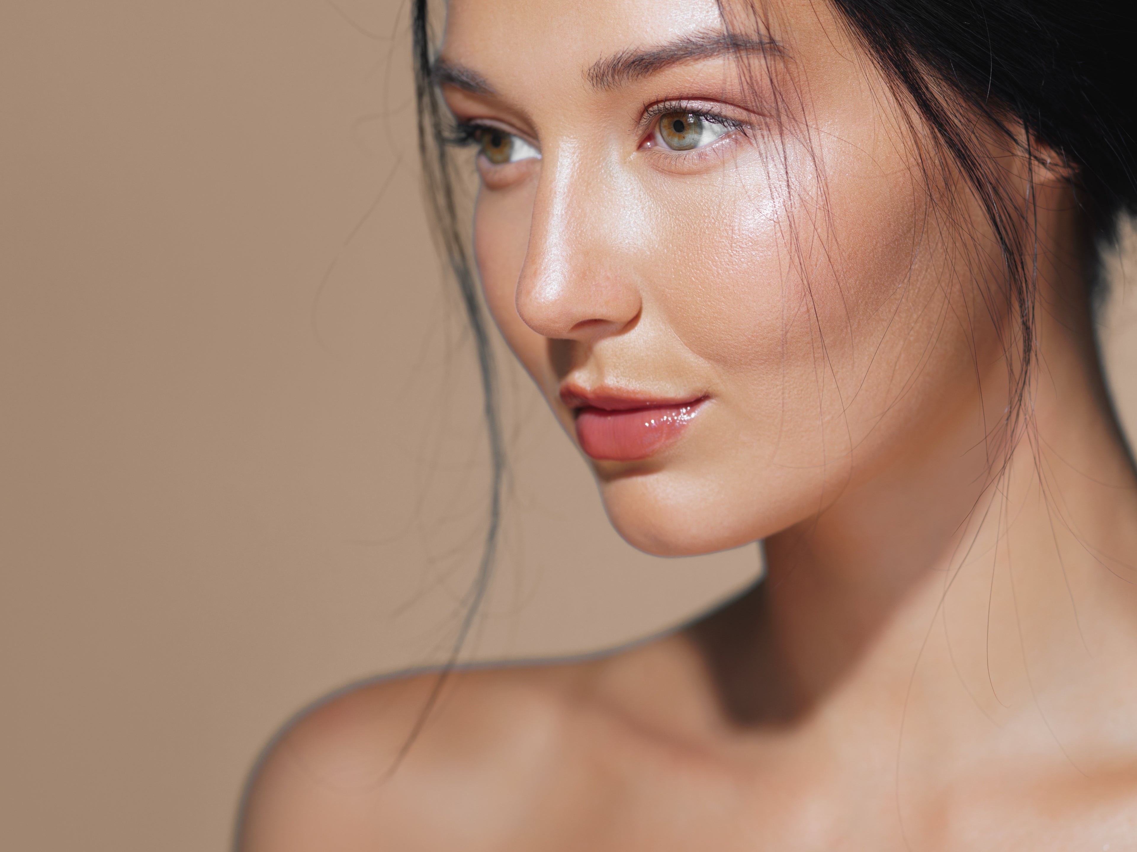 Close-up portrait of a woman with smooth, radiant skin and subtle makeup, shown against a soft beige studio background.