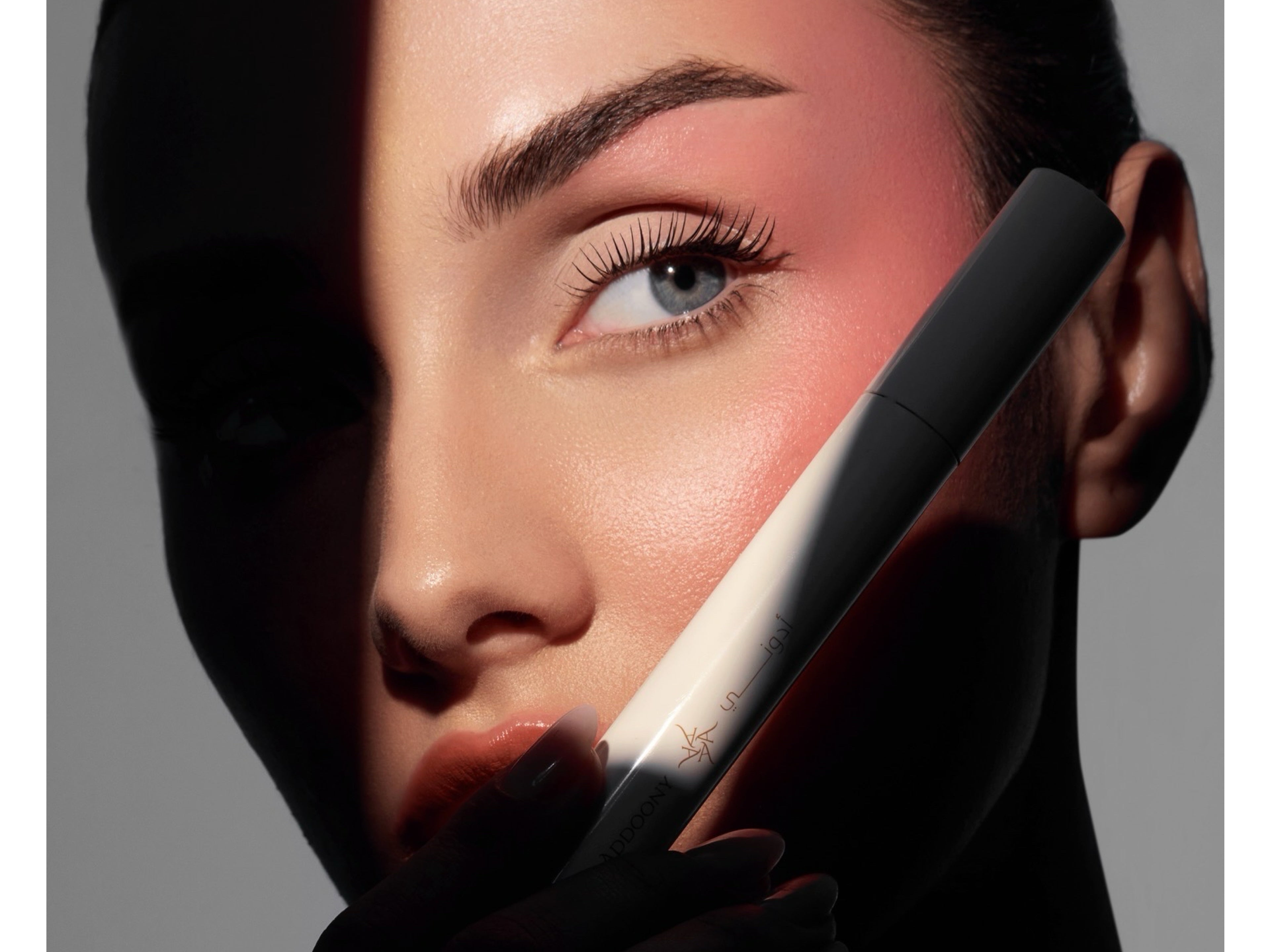 A close-up beauty shot of a model's face split by dramatic lighting, holding a black cosmetic tube against her cheek. The model has pink blush and is wearing a black glove, set against a dark gray background.
