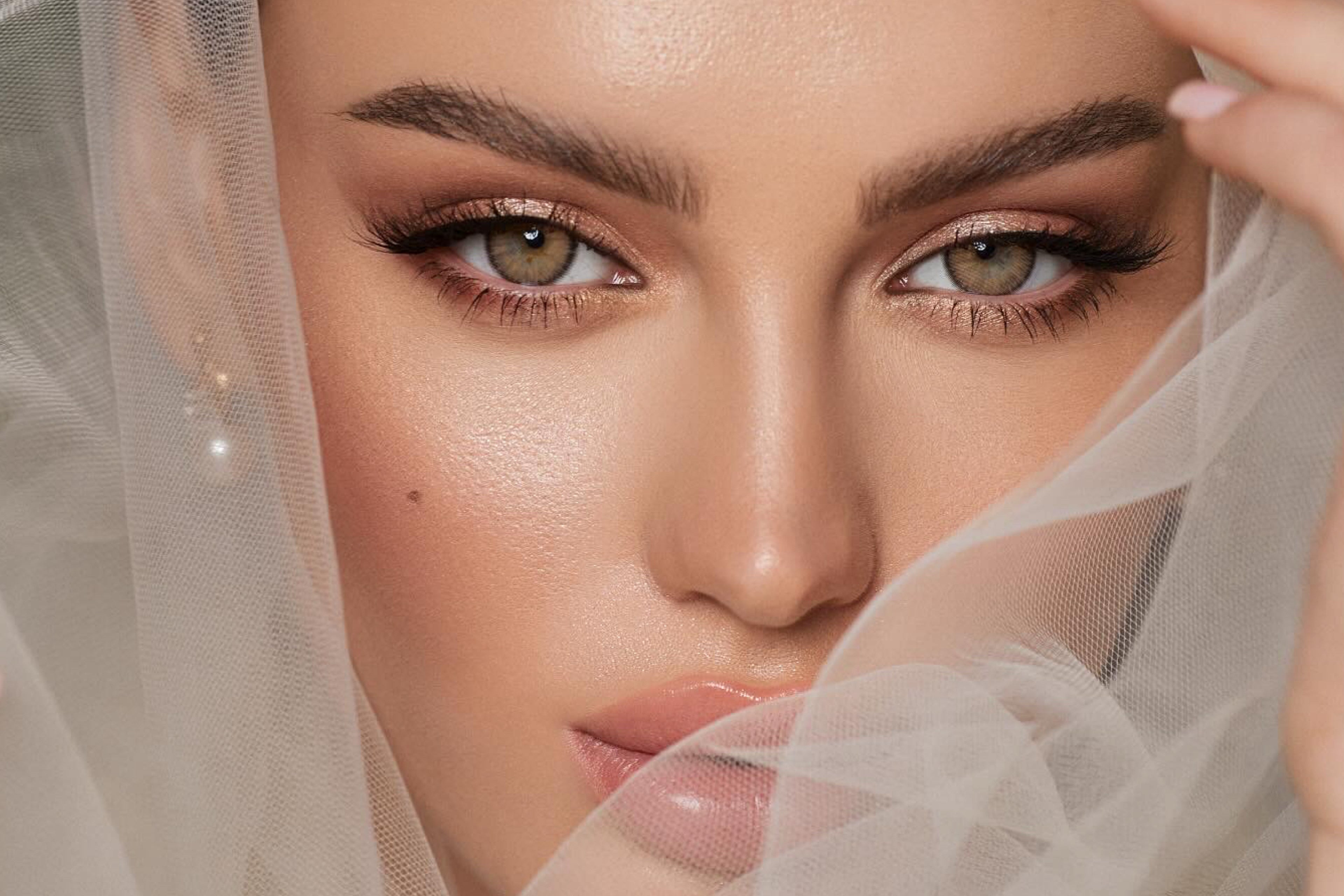A close-up portrait of a woman with bridal makeup and a sheer white veil, showing detailed eyes, brows, and glossy lips.