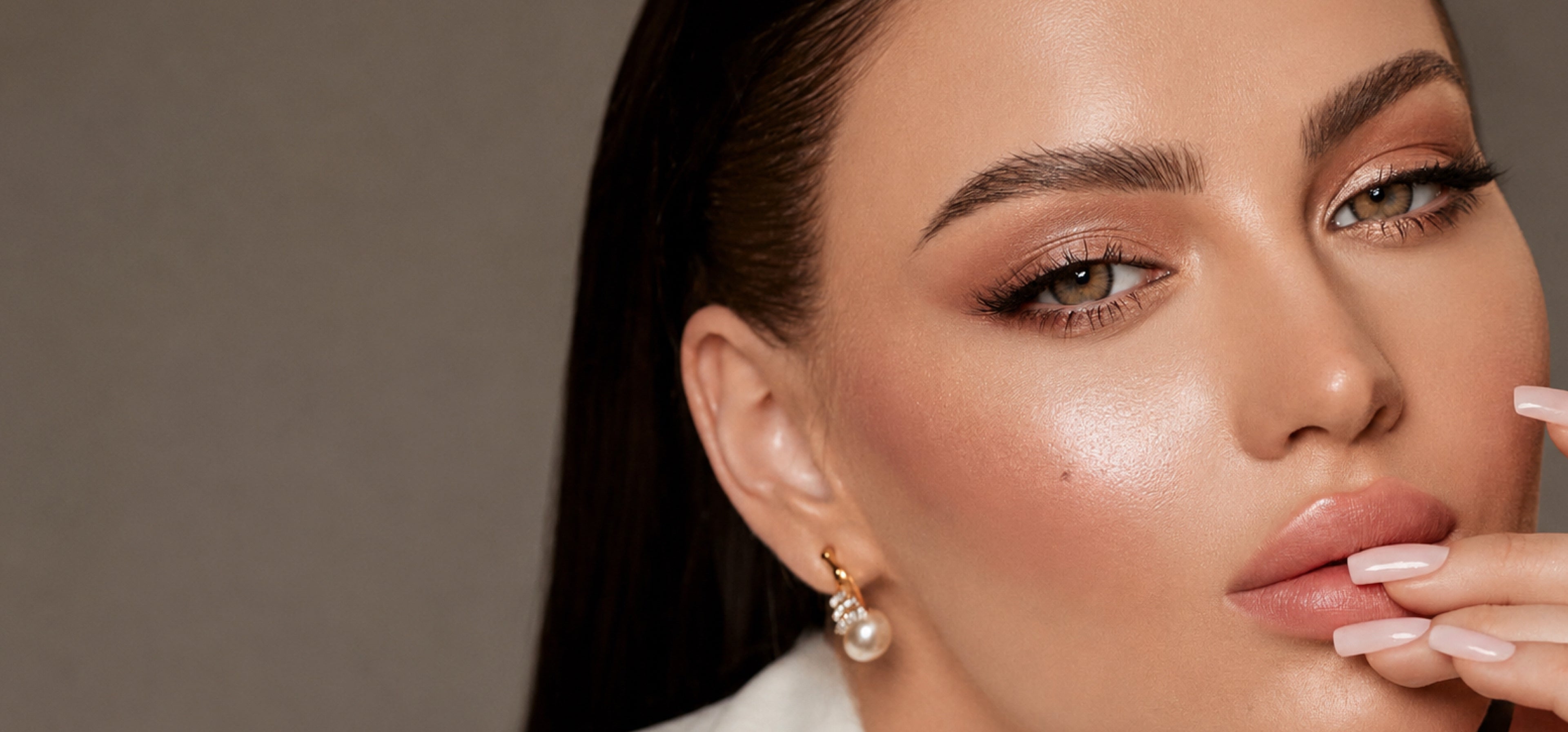 Close-up of a woman with dramatic eye makeup and a pearl drop earring, shown against a neutral gray background.