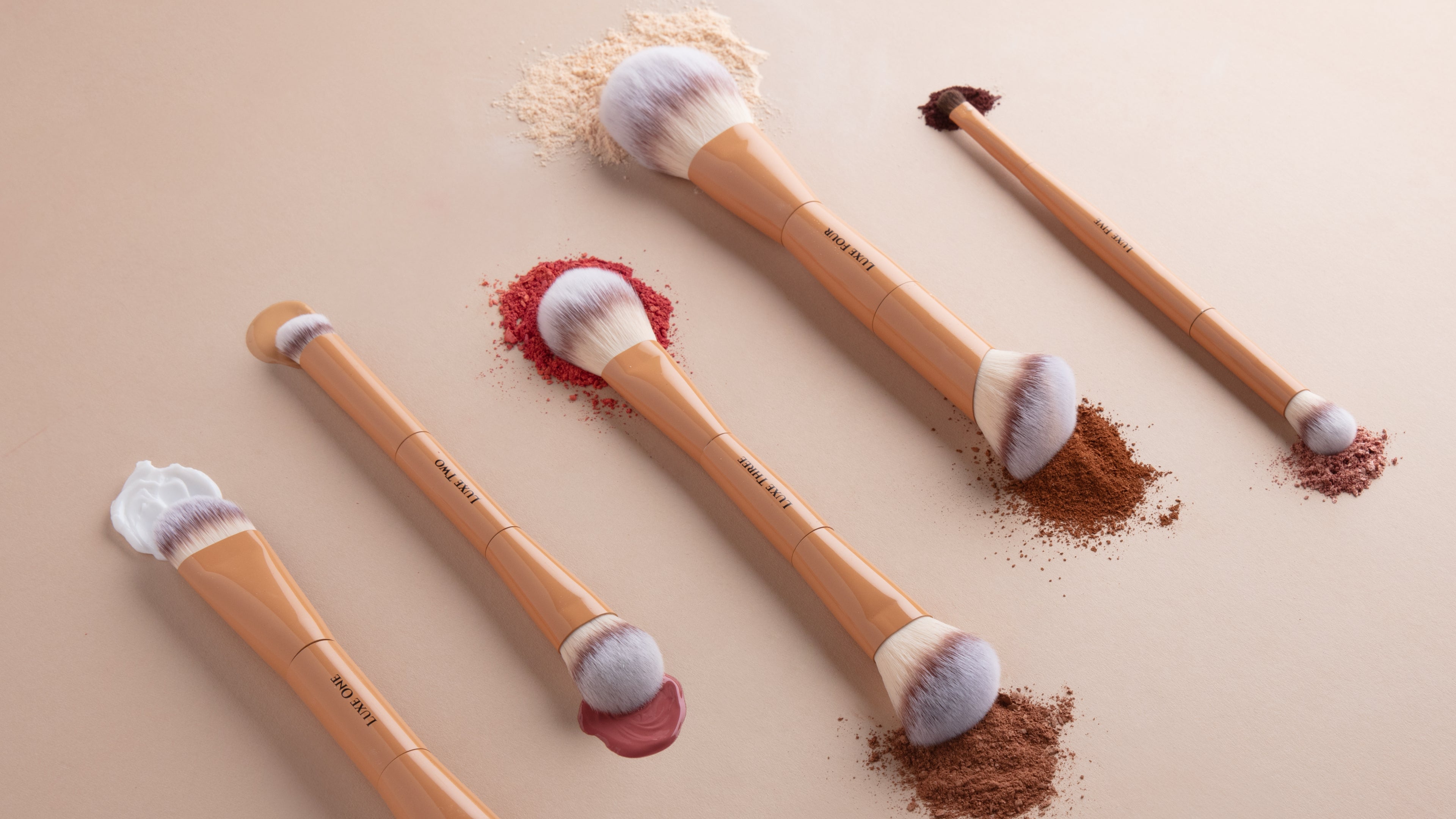 Four makeup brushes with light wood handles and white bristles are arranged on a warm beige background, each placed over a small pile of cosmetic powder in beige, red, brown, and mauve tones.