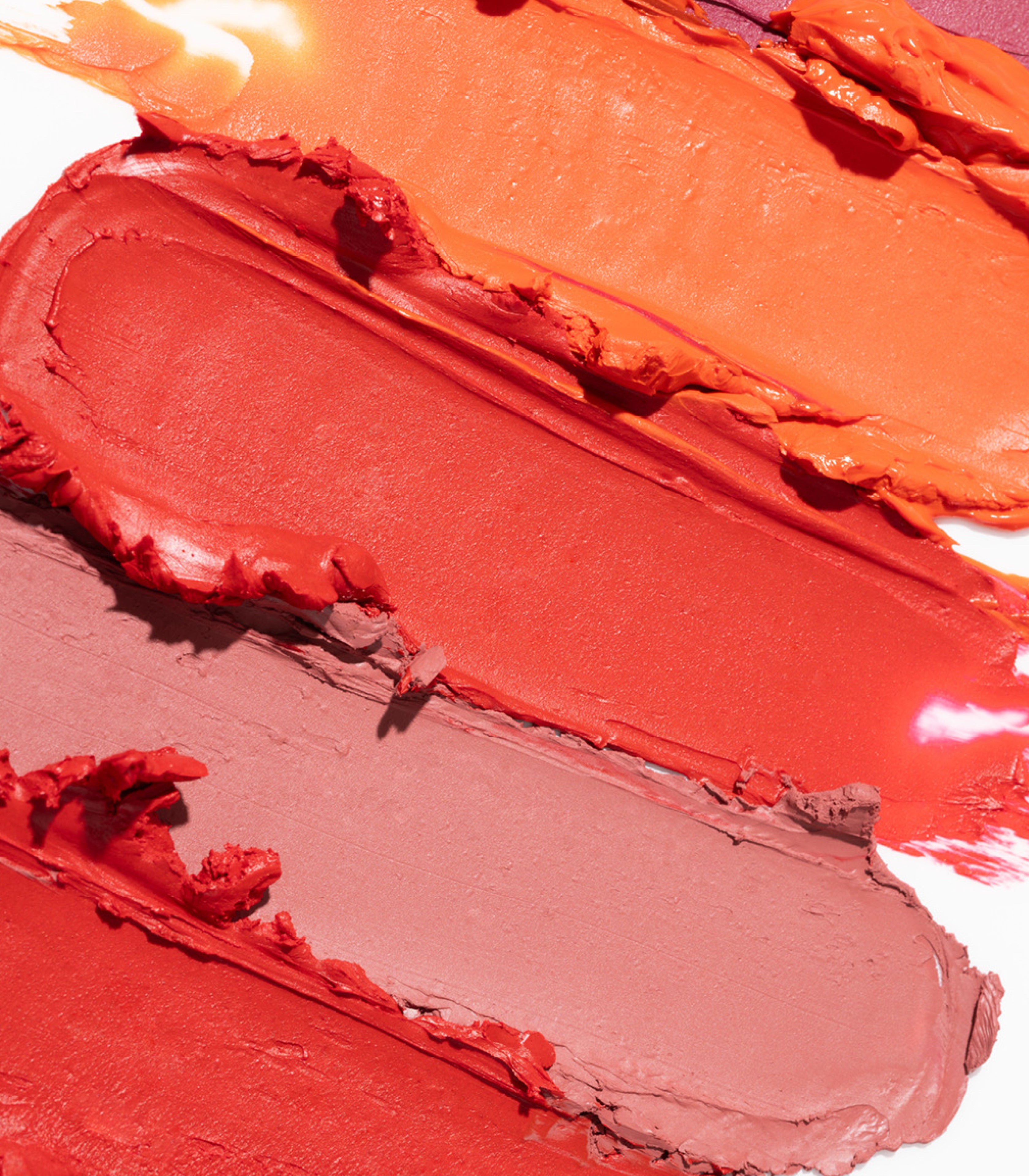 Close-up of thick, textured lipstick or cream makeup swatches in warm shades of coral, orange, red, and muted mauve on a white background.