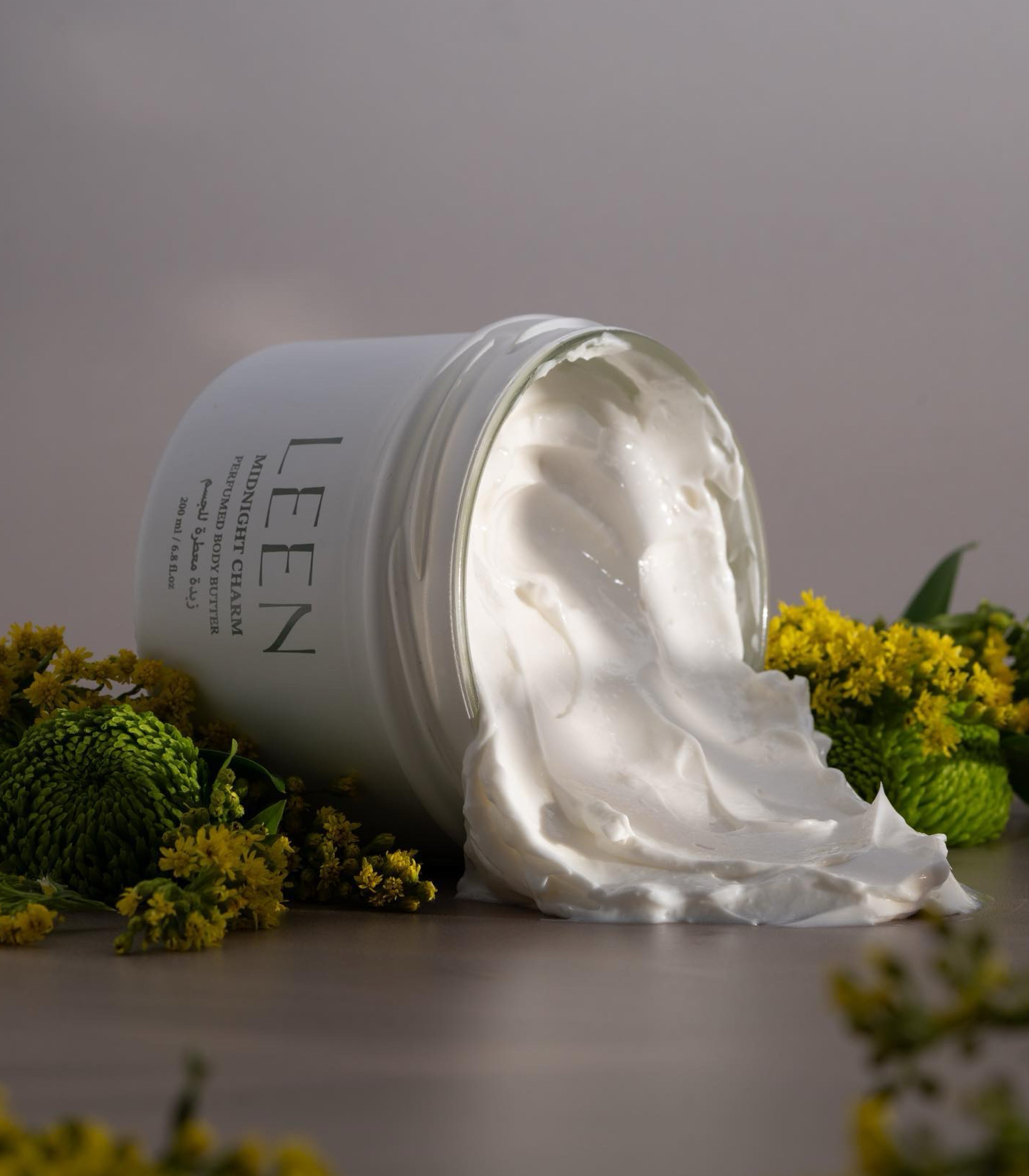 A white cosmetic jar labeled LEEN Moisturizing Cream, tipped over with thick white cream spilling out onto a surface, surrounded by small yellow flowers and green leaves against a soft neutral background.