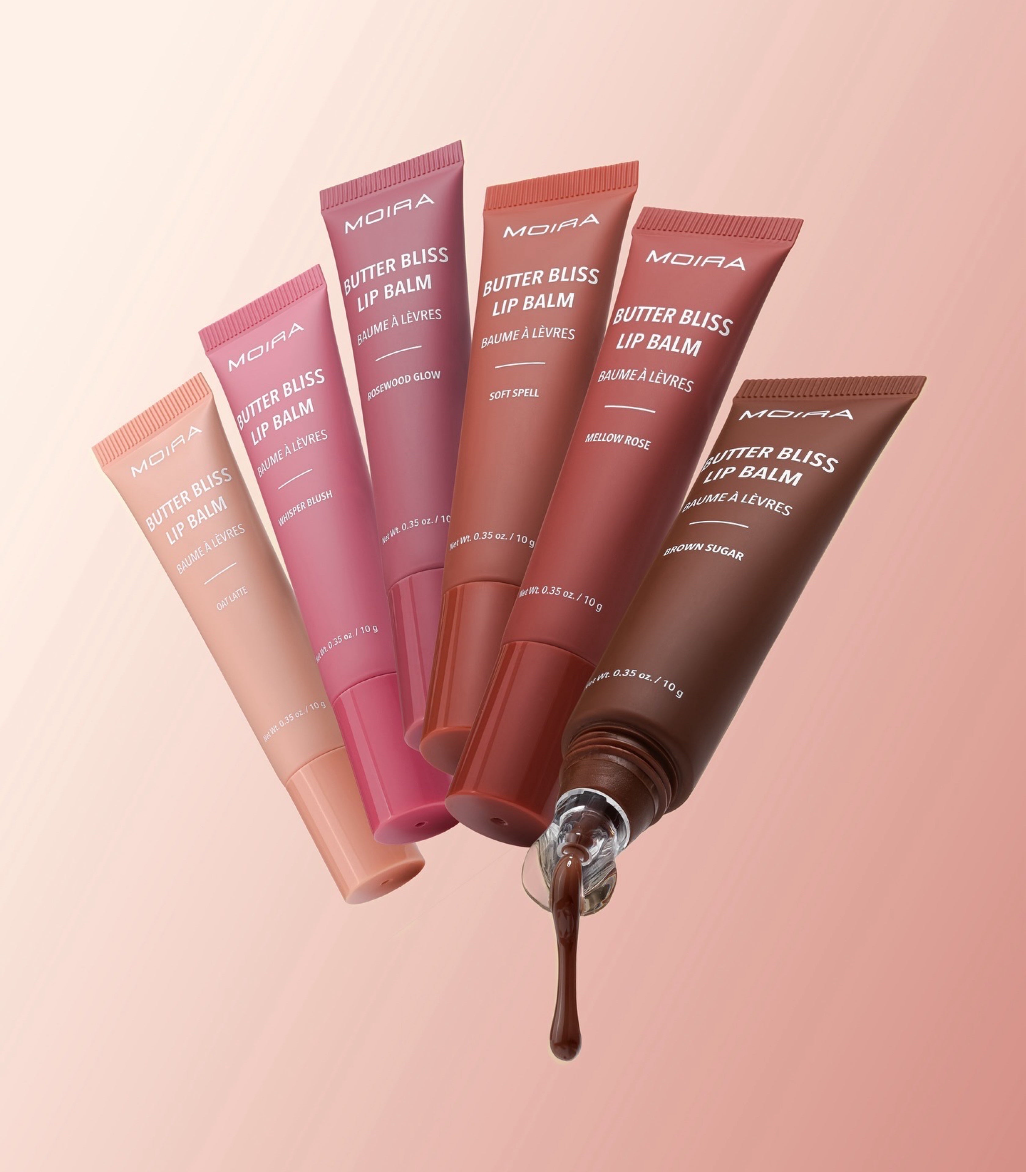 Six MOIRA Butter Bliss Lip Balm tubes in assorted shades from peach to deep brown, arranged in a fan shape with one tube open and a glossy balm drip dripping out, on a soft pink gradient background.
