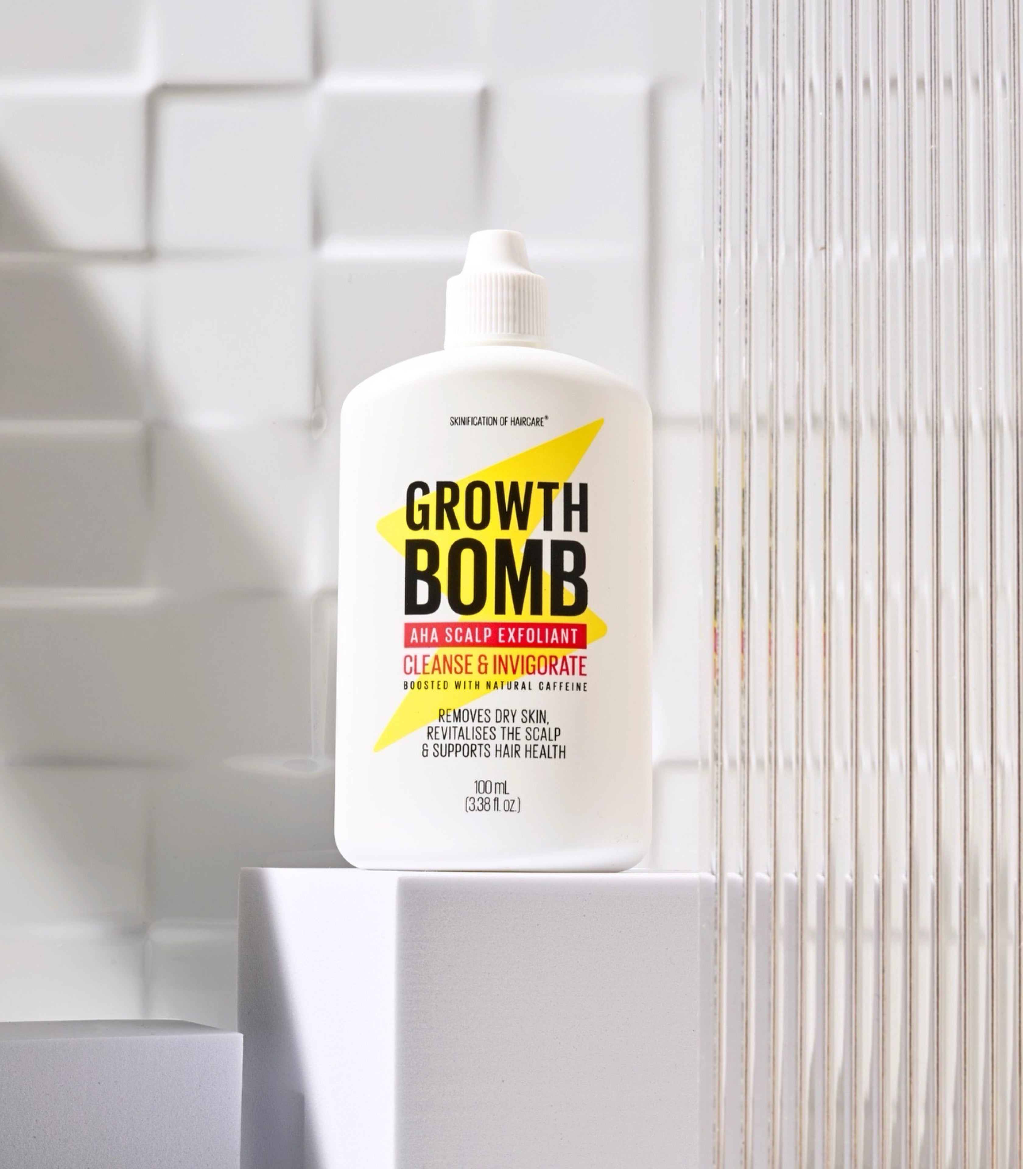 A white bottle of Growth Bomb Cleansing Balm with a yellow and black label, displayed on a white pedestal against a white tiled background.