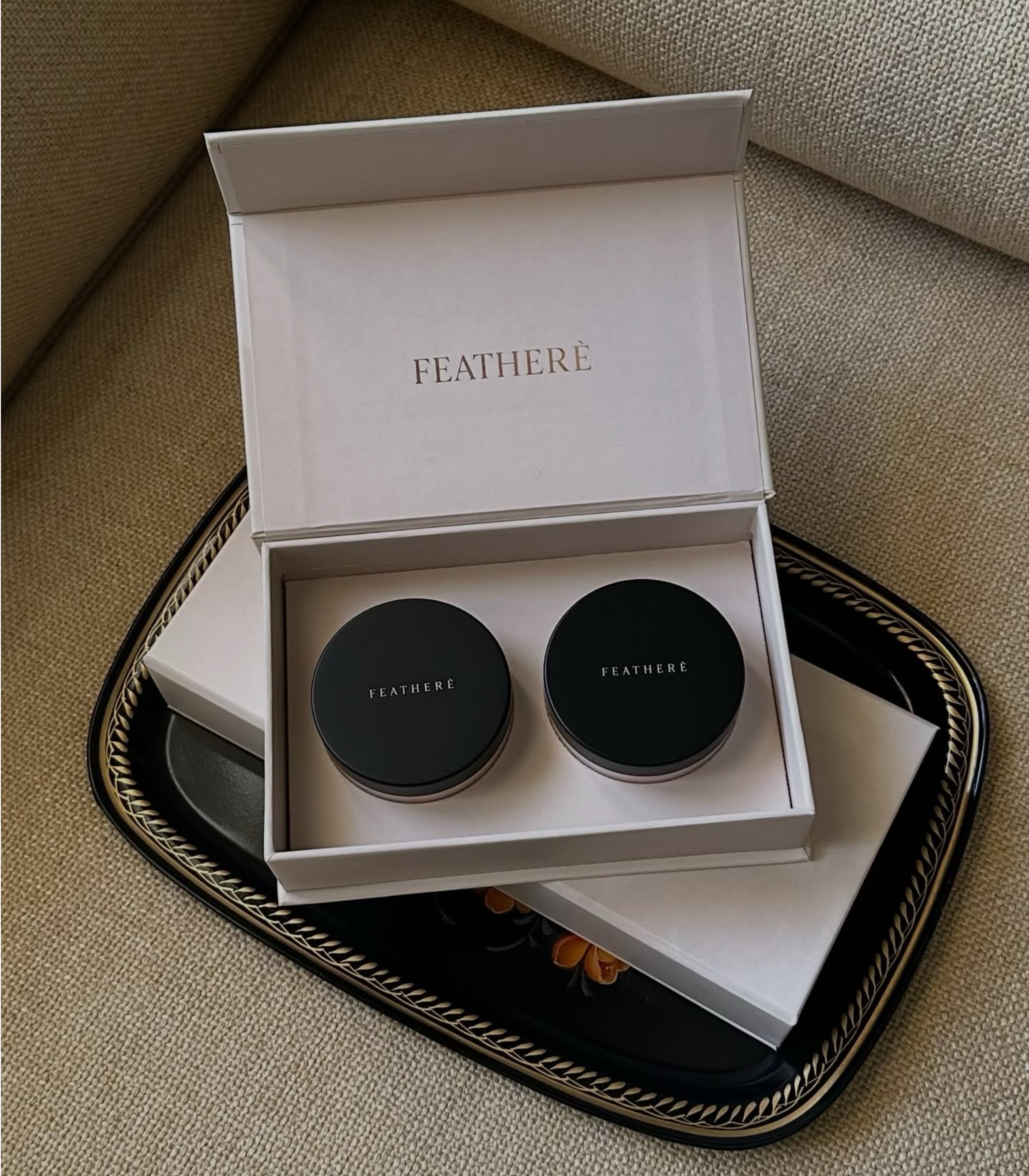 An open white FEATHERE gift box containing two round black cosmetic jars, placed on a black decorative tray over a beige textured fabric surface.