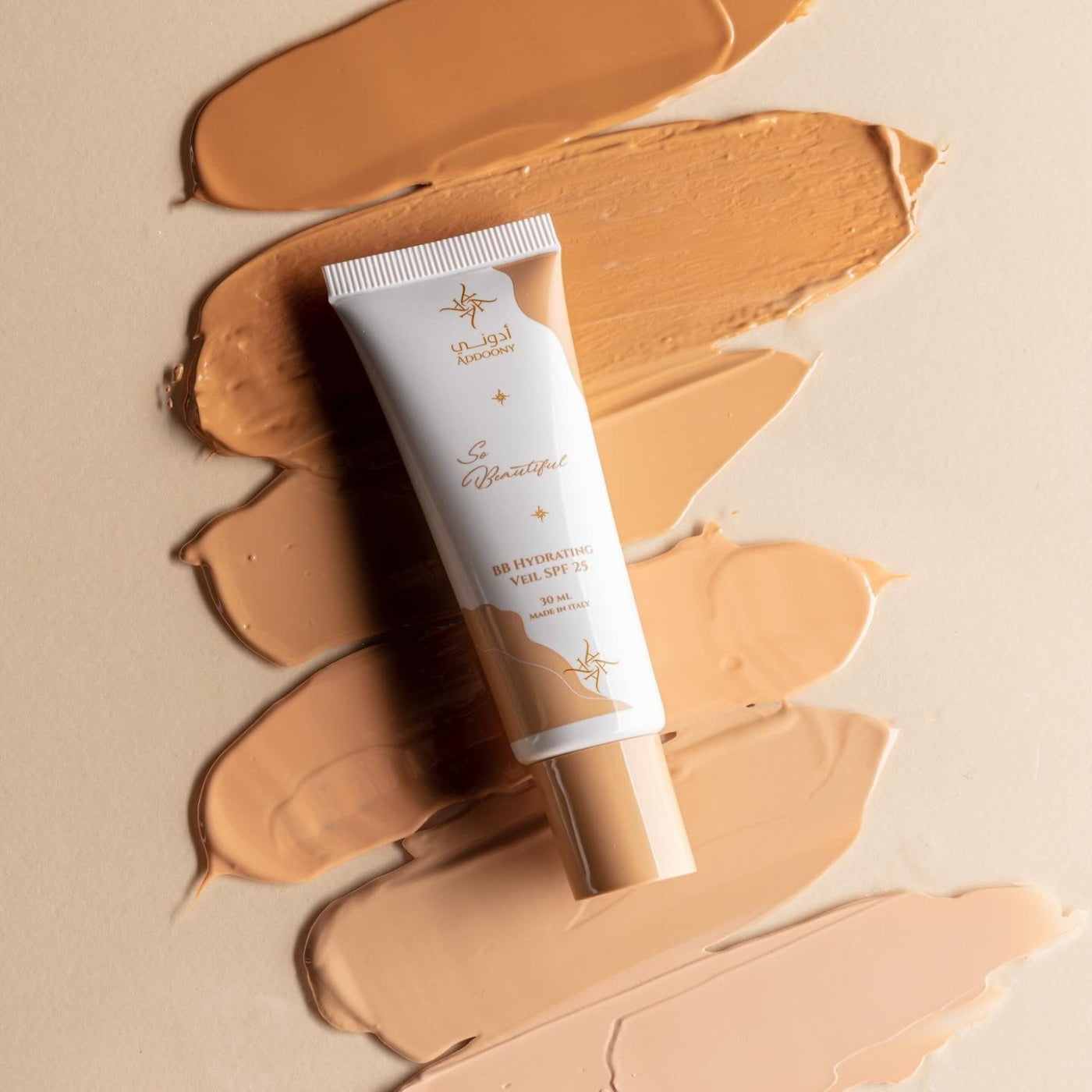 A squeeze tube of Hydrating Milk SPF 30 by Addoony Beauty, placed diagonally over several textured swatches of liquid foundation on a warm beige background.