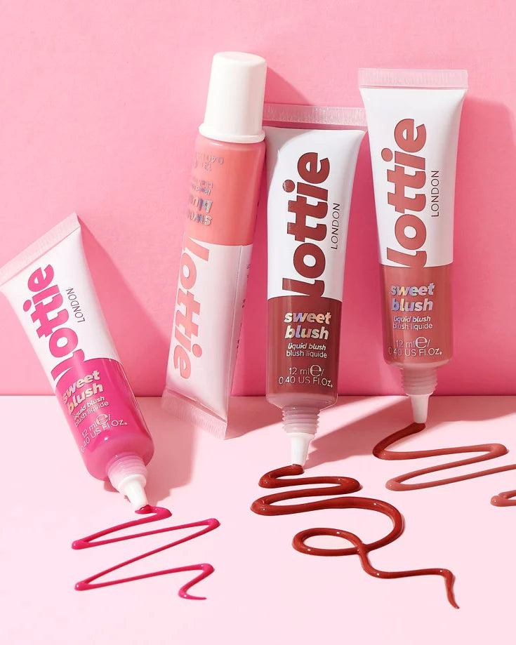 Four tubes of Lottie London Sweet Blush lip color arranged on a pink background, with pink, coral, and terracotta shades shown both in the tubes and as cosmetic swatches on the surface.