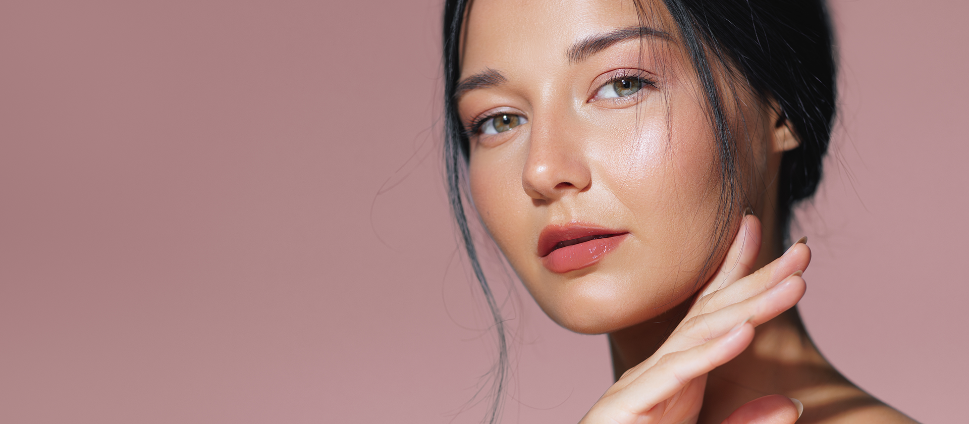 A close-up portrait of a woman with smooth, radiant skin and soft makeup, posing against a solid dusty pink background.