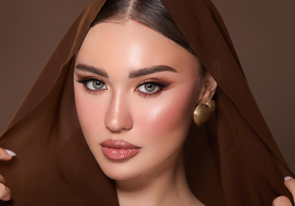 A close-up portrait of a woman wearing a brown headscarf or veil, with her face partially obscured by a large rectangular blur block. She has on dramatic eye makeup and a gold earring, set against a solid brown studio background.