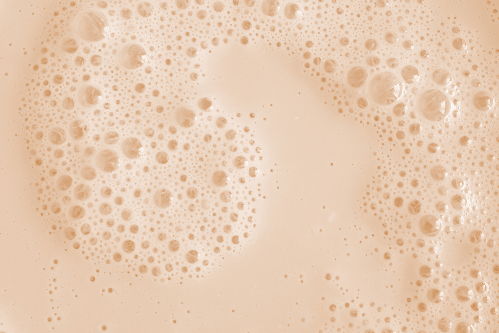 A close-up texture shot of a light beige liquid with dense, fine bubbles and larger bubbles clustered around the edges, creating a soft, creamy surface.